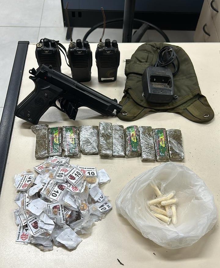 Material apreendido sobre a mesa na delegacia. Uma pistola de air soft, três rádios transmissores e uma base para carregamento, além de cocaína crack e maconha.